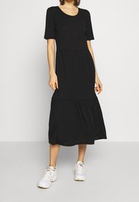 Kobieta w czarnej, krótkosleeve midi sukience i białych sneakersach, stojąca na tle jasnego, jednolitego tła.