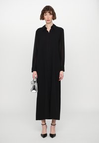 Vestido negro de manga larga con cuello y detalle de pliegue frontal, combinado con un bolso transparente y tacones negros de punta. Textura de tela suave.