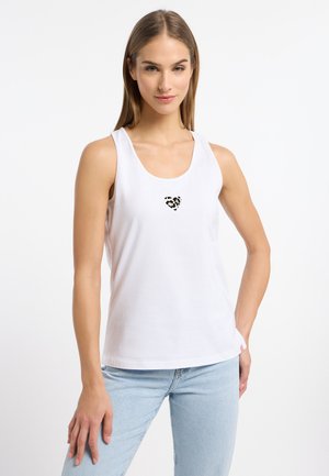 Kobieta ubrana w biały, bezrękawnik z małym sercem w panterkę na środku, w połączeniu z jasnoniebieskimi dżinsami, stojąca na białym tle.