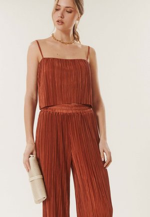 Femme portant un caraco plissé couleur rouille et un pantalon, tenant une pochette beige roulée, sur un fond clair uni.
