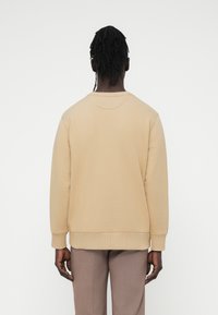 Μπεζ φούτερ με στρογγυλή λαιμόκοψη, ριμπ μανσέτες και τελείωμα, με ομαλή υφή. Φορεμένο με ανοιχτό καφέ παντελόνι.