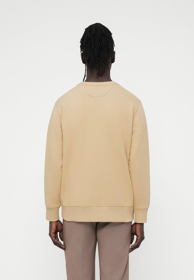 Sweat-shirt beige à col rond, poignets et ourlet côtelés, avec une texture lisse. Porté avec un pantalon marron clair.