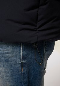Marinblå puffjacka med slät yta, snörningsdetalj vid fållen, tillsammans med bleka blå denimjeans.
