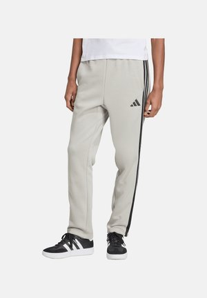 Dolna część ciała osoby noszącej jasnoszare spodnie dresowe Adidas z czarnymi bocznymi paskami oraz czarne buty sportowe Adidas z białą podeszwą.