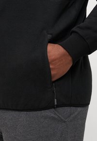Crna flis majica s džepom na zatvarač i rebrastim manžetama, s mekanom teksturom i opuštenim krojem, u kombinaciji s sivim trenerkama.