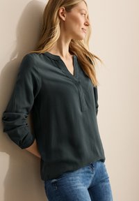 Donkergroene, gestructureerde blouse met een V-hals en opgerolde mouwen, gecombineerd met blauwe denim jeans. Staand tegen een neutrale muur.