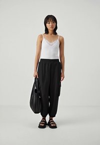 Black cargo pants with pockets, white tank top with lace trim, and black slide sandals; model holds a black textured tote bag.