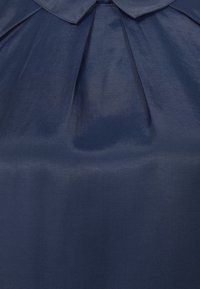 Marinblå blus med krage, plisserad fram och slät yta, tillverkad av lättviktig tyg.