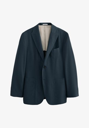 Man in een marineblauwe blazer, gestreept overhemd, olijfgroene broek en zwarte loafers leunt tegen een beige muur en kijkt opzij.