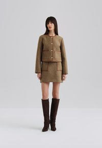 Giacca in tweed marrone con bottoni dorati e rifiniture marroni, abbinata a una gonna coordinata con tasche. Stivali lunghi in suede marrone completano l'outfit.