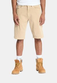 Pantaloni cargo beige con due tasche frontali, indossati con stivali impermeabili color cammello. Calzini bianchi con logo visibile in cima. Design semplice e funzionale.