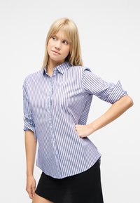 Γυναίκα με ξανθά μαλλιά που φοράει μπλε και λευκό πουκάμισο με κάθετες ρίγες και μαύρη φούστα, ποζάροντας με το ένα χέρι στη μέση, κοιτάζοντας προς τα αριστερά.