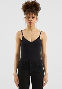 Vrouw met krullend haar in een hoge bun draagt een zwart spaghettitop en zwarte jeans, staand tegen een effen lichte achtergrond.