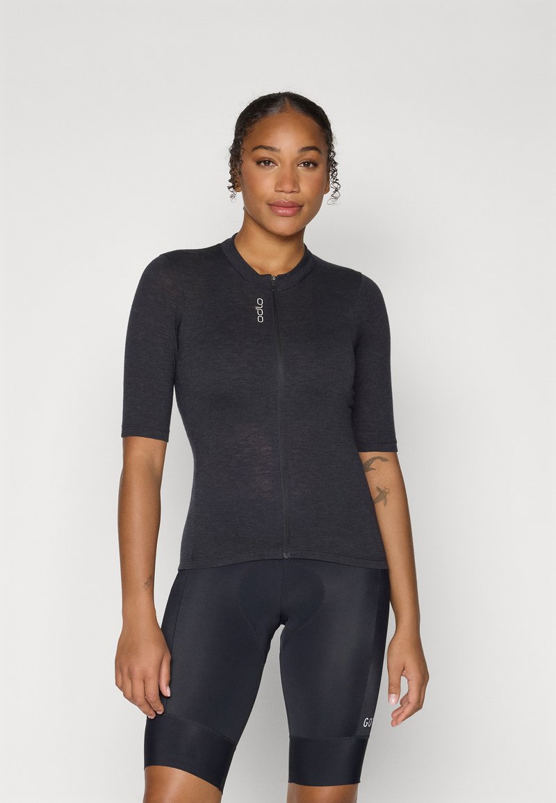 Haut zippé à manches courtes en tissu gris foncé, avec un col rond et une coupe ajustée. Porté avec un short noir, mettant en avant une surface texturée.