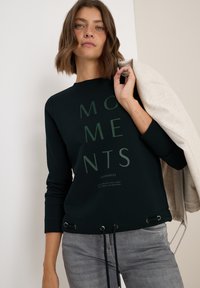 Zwarte sweatshirt met de tekst "MOMENTS" in donkergroen, voorzien van een trekkoord aan de onderkant, ronde halslijn en lange mouwen. Grijze jeans en een lichte jas.