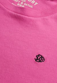 Lyserød bomulds t-shirt med rund hals, med sort broderet logo på fronten nær halsudskæringen. Blød tekstur og sømfri finish.