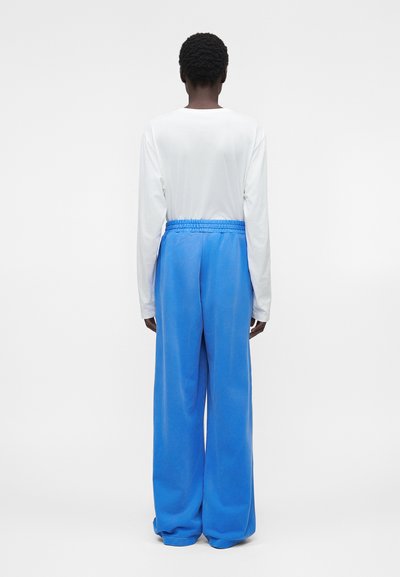 Άτομο με κοντά σγουρά μαλλιά, που φοράει λευκό μακρυμάνικο πουκάμισο και φαρδιά μπλε παντελόνια, στέκεται με την πλάτη προς απλό φόντο.