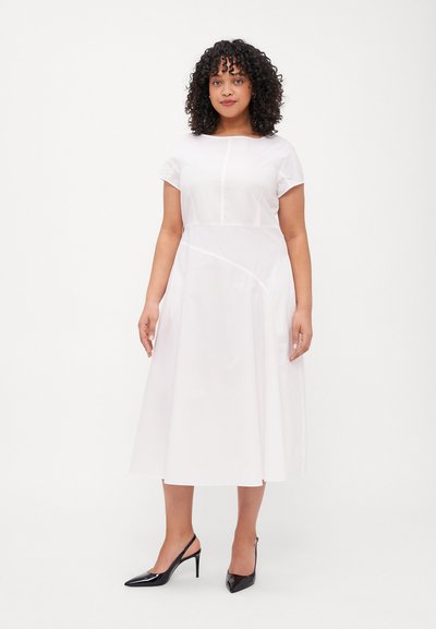 Robe blanche à manches courtes, décolleté rond et silhouette en A. Présente des détails à coutures courbes et une texture lisse. Associée à des talons noirs.