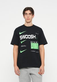 Črna majica s kratkimi rokavi ima bold belega "SWOOSH" napisa in zeleni poudarki. Oblika vključuje Nike logotipe in mrežasti vzorec. Narejena iz bombažne tkanine.