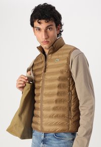 Vesta puf de culoare maronie cu un model cu quilt orizontal, guler înalt și buzunare cu fermoar. Logo pe pieptul stâng. Purtați-o peste o cămașă bej cu mânecă lungă.