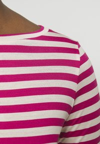 Nahaufnahme einer Person, die ein pink-weiß gestreiftes Hemd mit rundem Ausschnitt vor einem einfarbigen Hintergrund trägt.