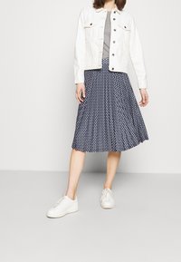 Bílá džínová bunda, šedá košile, tmavě modrá plisovaná sukně s bílými puntíky a bílé tenisky. Hladká textura a volnočasový design.