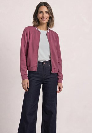 Kvinde smilende iført en pink lynlåsjakke, hvid t-shirt og mørke, vide jeans mod en ensfarvet lys baggrund.