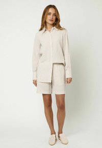 Camisa de botones a rayas beige con mangas largas, combinada con pantalones cortos a juego. La tela es texturizada y ligera, complementada con calzado casual.