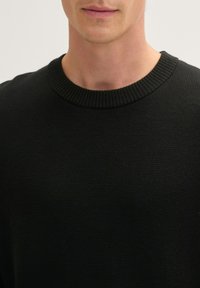 Schwarzer Strickpullover mit einem gerippten Rundhalsausschnitt und strukturierter Oberfläche. Nahaufnahme, die sich auf die Details des Materials und des Ausschnitts konzentriert.
