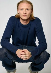 Pull en tricot bleu marine avec des détails côtelés aux épaules, associé à un jean baggy en denim foncé. Le modèle est accroupi, montrant ses mains tatouées.