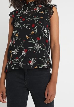 Mouwloze zwarte blouse met gerimpelde kraag en zoom, met rode, witte en gele bloemenpatronen, gedragen met zwarte broek.