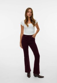 Mujer con cabello largo castaño, vistiendo una blusa blanca ajustada de manga corta, pantalones de pana burdeos y botas oscuras de punta, de pie con la mano en el bolsillo.