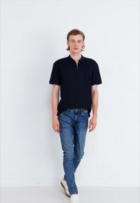Camisa polo azul marino con cuello con cremallera, combinada con jeans ajustados azul claro y zapatillas blancas. Tejido suave sin patrones ni acentos visibles.