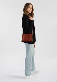 Borsa in pelle marrone con motivo a griglia, di media grandezza, con tracolla regolabile e dettagli in hardware argentato, abbinata a un blazer nero e jeans chiari.