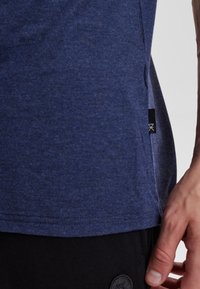 Námořnická modrá bavlněná tričko s texturovaným povrchem, bočním rozparkem a malou černou štítkou na lemu. Navrženo pro pohodlí a každodenní nošení.