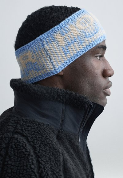 Ung svart man i profil, iförd ett ljusblått och beigefärgat stickat pannband och en svart texturerad zip-up-jacka med hög krage