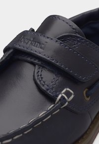 Μπλε ναυτικό δερμάτινο παπούτσι σκαφών με ραμμένες λεπτομέρειες, velcro με ανάγλυφο το όνομα «Timberland» και ορατές οπές με κορδόνια.