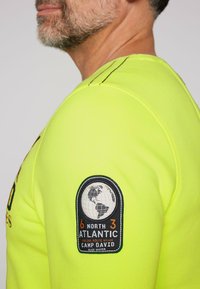 Sudadera amarilla neón con un parche redondo que muestra un globo, costuras negras y detalles de texto en el hombro izquierdo. Textura suave, diseño casual.