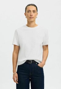 Femme aux cheveux foncés attachés, portant une chemise blanche à manches courtes, un jean foncé avec une ceinture noire, ainsi que des boucles d'oreilles créoles argentées et un bracelet.