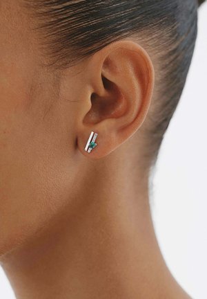 Primer plano de la oreja de una persona con un pendiente plateado que tiene dos barras verticales y una pequeña gema verde.