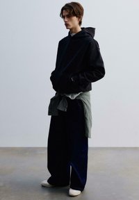 Giovane uomo che indossa un hoodie nero, pantaloni larghi scuri, sneakers bianche e una camicia a quadri annodata in vita, in piedi su un pavimento semplice.