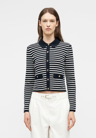 Γυναίκα που φοράει μαύρη και λευκή μακρυμάνικη ζακέτα με οριζόντιες ρίγες και χρυσά κουμπιά, καθώς και λευκό παντελόνι ψηλόμεσο με ζώνη.