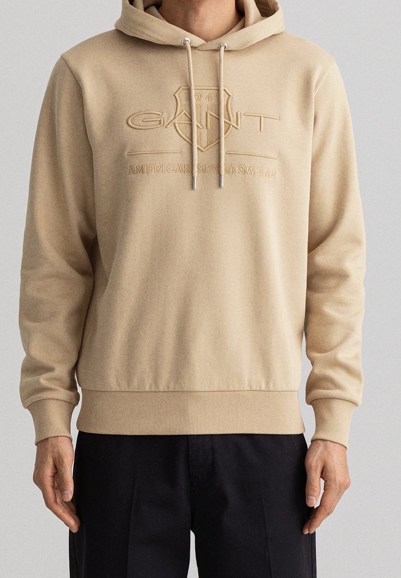Beige Kapuzenpullover aus weichem Material, mit einem geprägten Logo und Kordeln mit silbernen Enden. Elastische Bündchen und Saum runden das Design ab.
