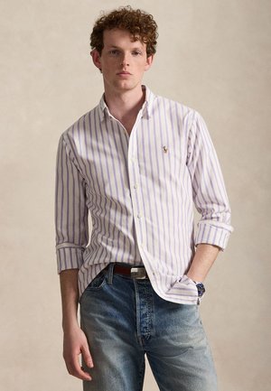 Nuori mies, jolla on kiharat hiukset, pukeutunut valkoiseen ja laventelin väriseen raidalliseen nappikauluspaitaan, sinisiin farkkuihin ja rannekelloon, seisoo tasaisen taustan edessä.