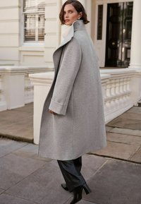 Cappotto di lana grigia con colletto alto, tasche laterali e maniche lunghe. Indossato sopra pantaloni in pelle nera e stivali con tacco, con una texture liscia.