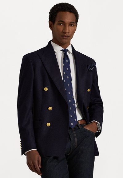 Ναυτικό διπλόστατο σακάκι με χρυσά κουμπιά, συνδυασμένο με πασέ κρουαζέ και μαντήλι τσέπης. Φοριέται πάνω από ένα λευκό πουκάμισο και σκούρα τζιν.