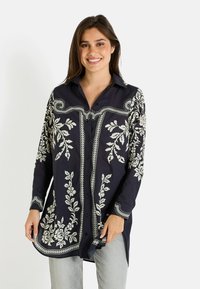 Camisa navy de botones con bordado floral blanco, de mangas largas y dobladillo curvado. Presenta bordes en contraste y cuello de tipo camisa.