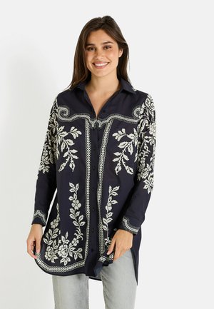 Camisa navy de botones con bordado floral blanco, de mangas largas y dobladillo curvado. Presenta bordes en contraste y cuello de tipo camisa.