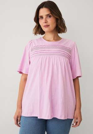 Woman with shoulder-length brown hair wearing a loose light pink short-sleeve top with lace detailing across the chest and blue jeans.