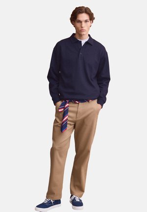 Giovane uomo con un maglione blu navy, pantaloni beige con cintura a righe annodata e sneakers blu, in piedi con le mani nelle tasche.